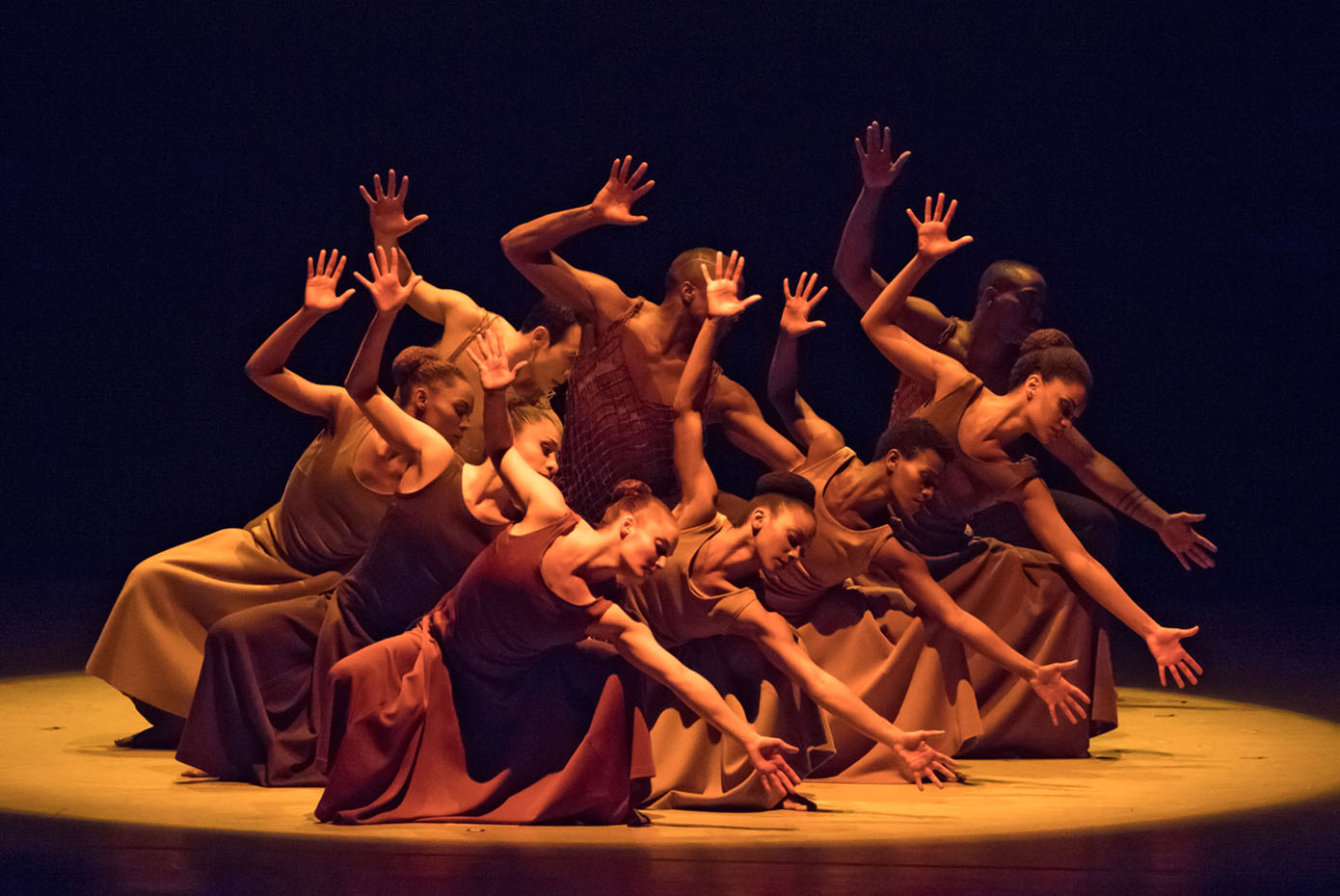 Alvin Ailey dancers in costume are in a spotlight on a stage.