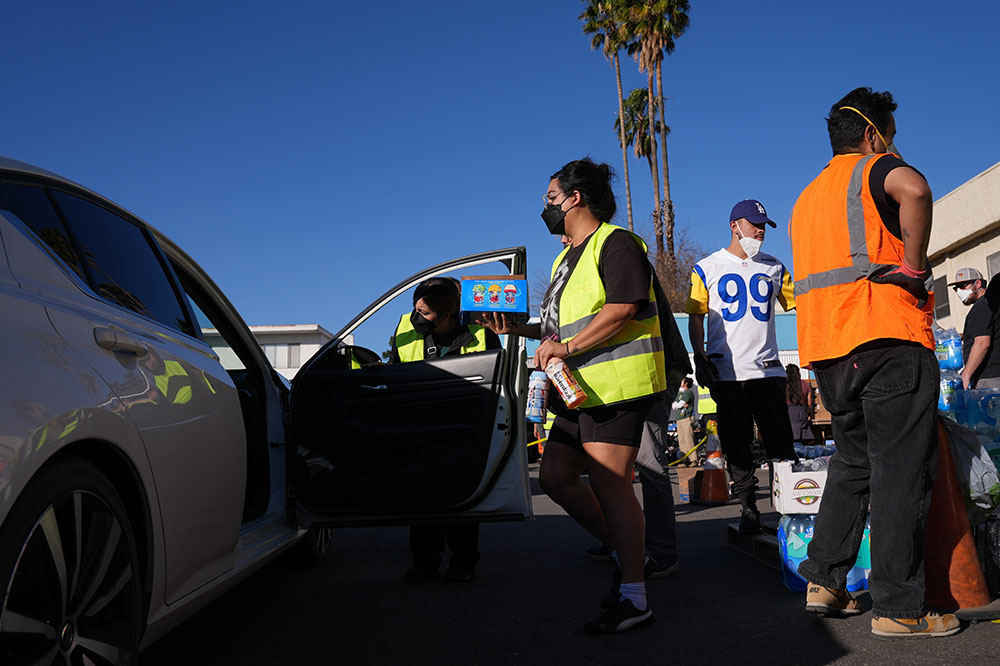 A woman wearing a neon vest gives bottled beverages and other supplies to an unseen person inside a car.