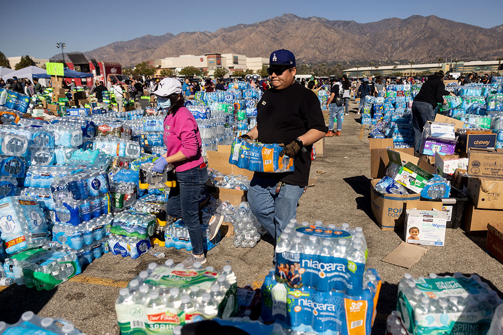 A man and a woman carry packages of bottled water amid piles of similar packages in an open lot.