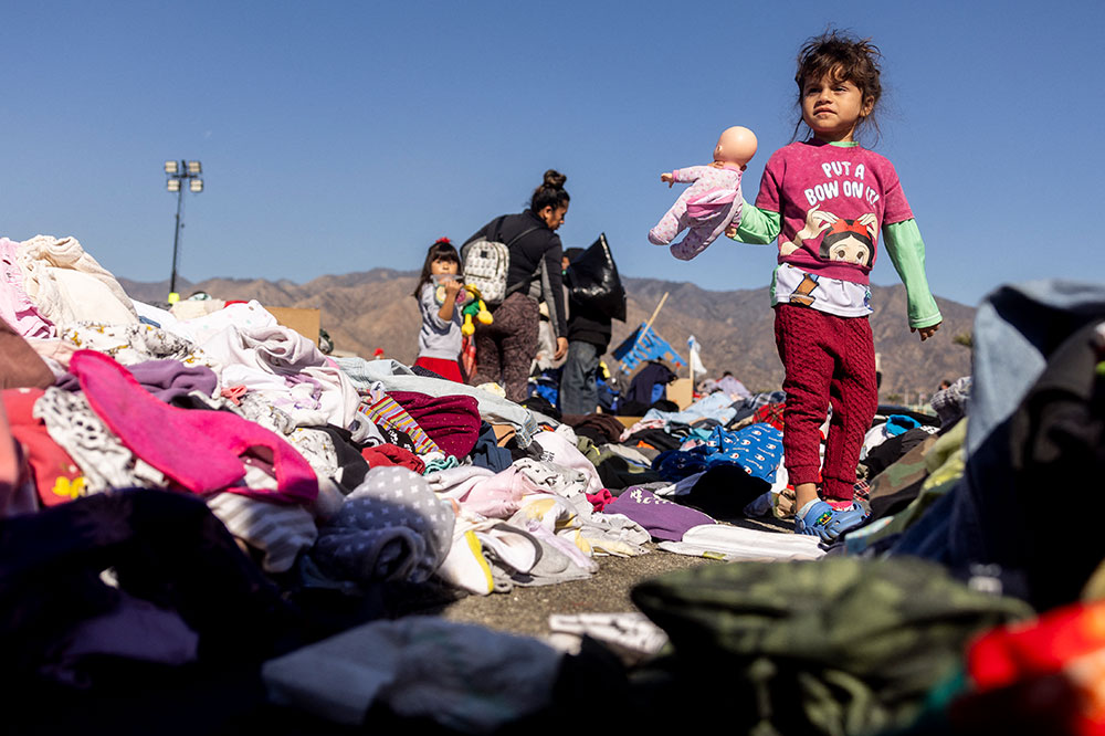 A child stands in the middle of a pile of donated clothing and holds up a doll.