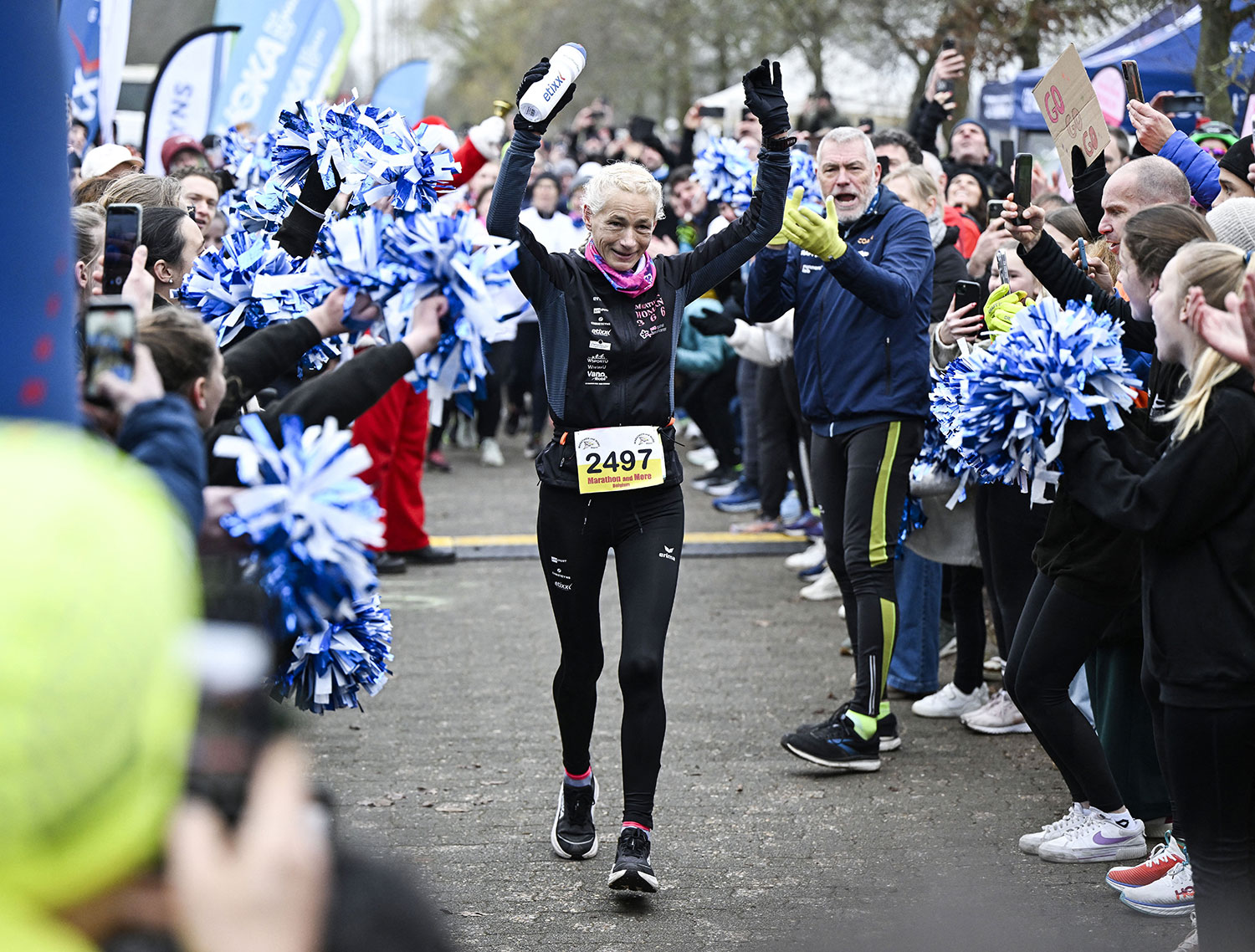 Hilde Dosogne wears running clothes and a number and has her arms up in triumph as she crosses a finish line surrounded by spectators.