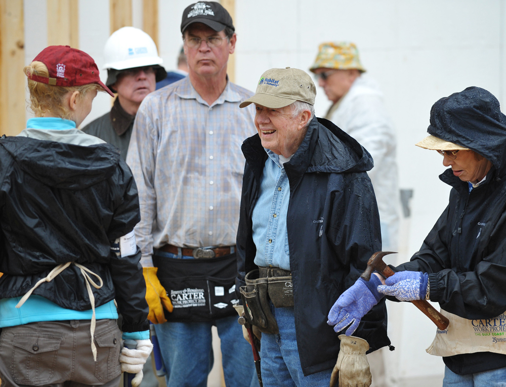 Older Jimmy and Rosalynn Carter wear jackets and toolbelts as they mingle with others at a construction site.