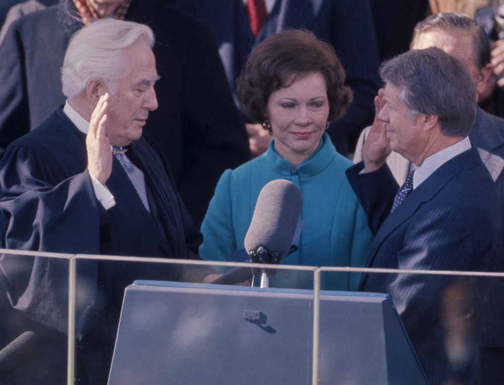 Jimmy Carter stands with his right hand raised. He is beside a podium and next to Rosalynn Carter and facing a judge.