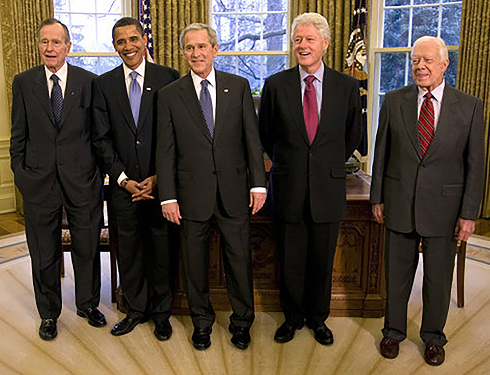 George Bush, Barack Obama, George W. Bush, Bill Clinton, and Jimmy Carter pose in front of the desk in the Oval Office.