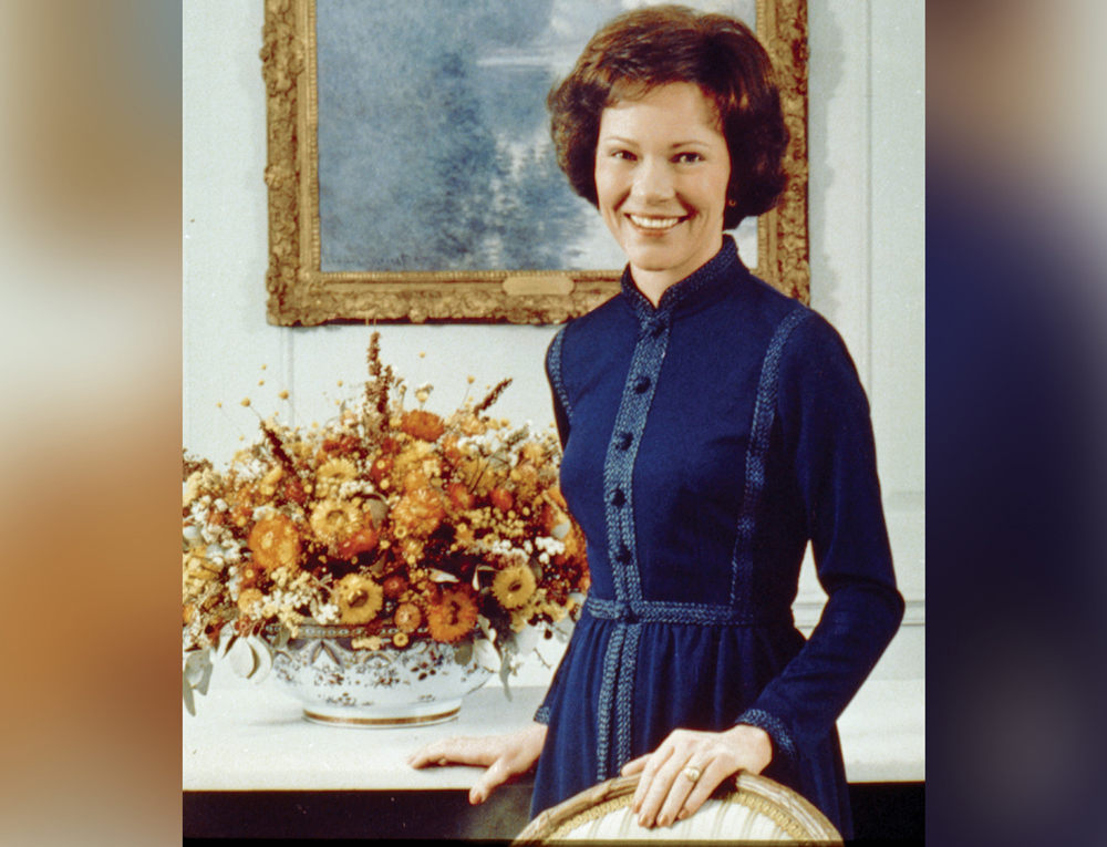 A standing Rosalynn Carter poses and smiles with her hand on a chair.