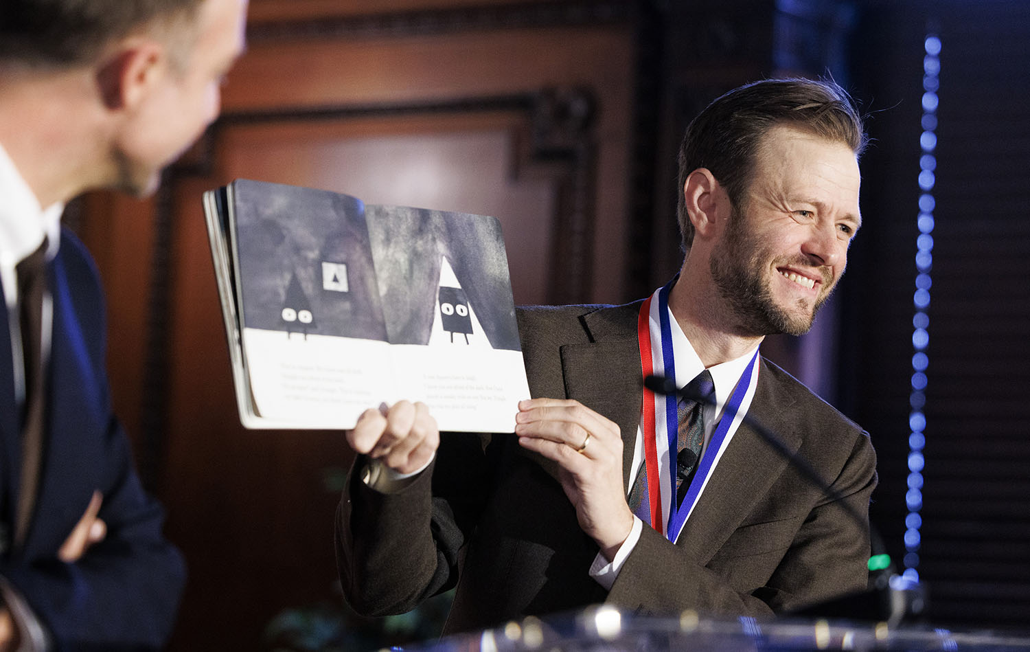 Mac Barnett smiles as he holds open his book, Triangle, to show the illustrations to an audience as Jon Klassen looks on.