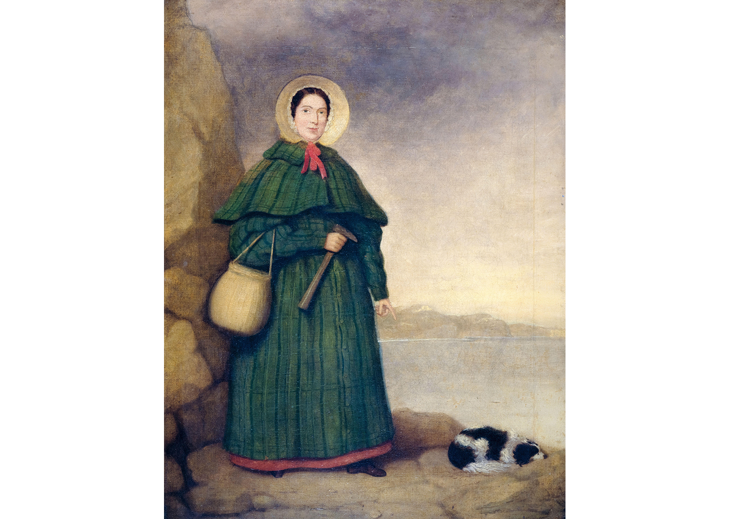 Mary Anning stands outdoors holding a basket and a tool as her dog lies next to her.