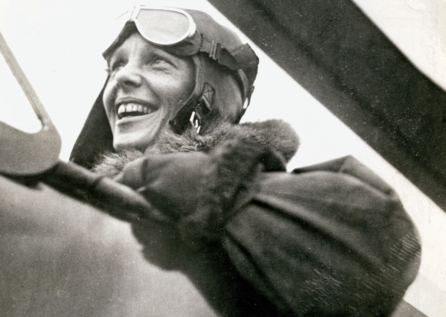 Amelia Earhart sits in the cockpit of an open-air plane wearing goggles and a helmet.