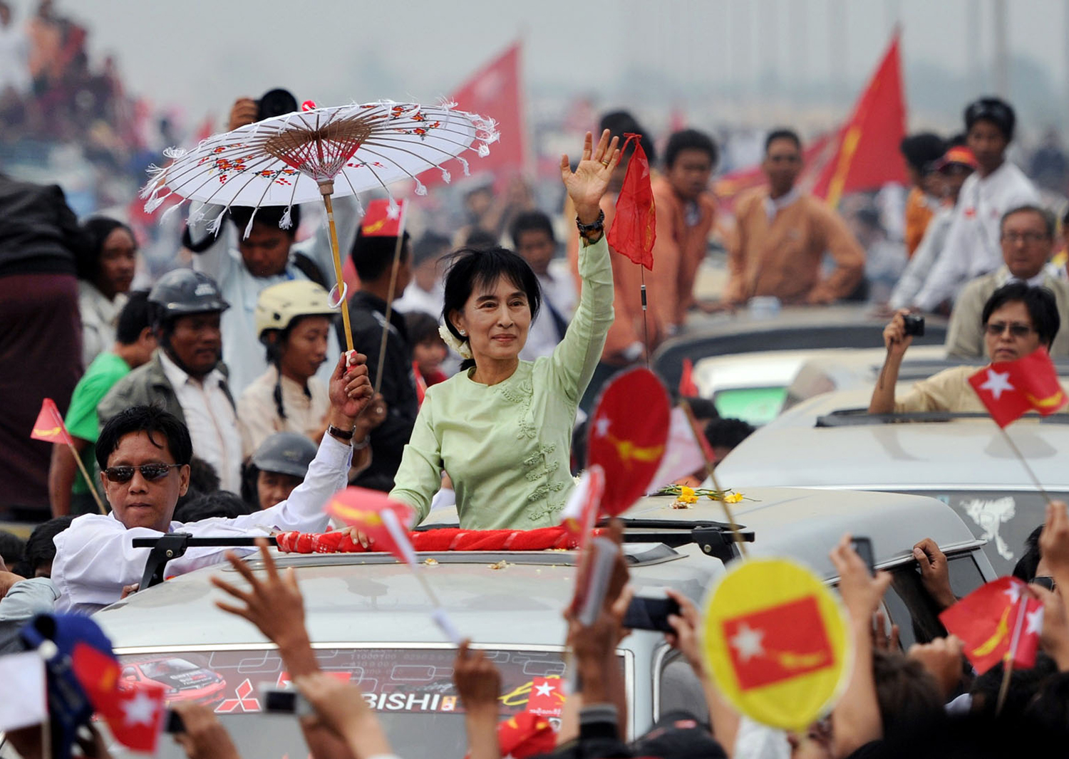 Aung San Suu Kyi stands in a car with the sunroof open and waves to a crowd during a procession