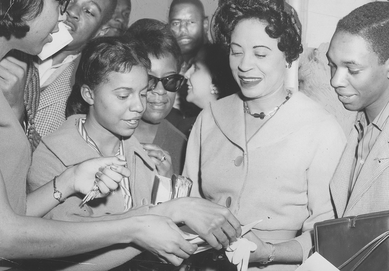 Daisy Bates smiles as she signs a piece of paper for a teen amid a crowd of other teens waiting for autographs.