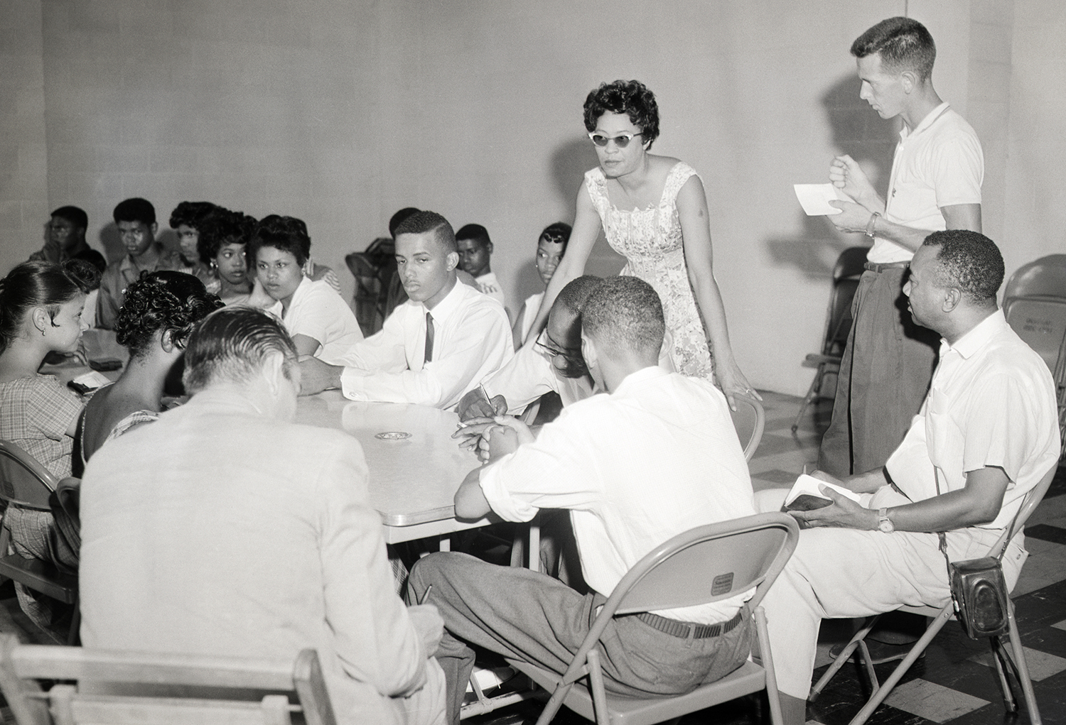 Daisy Bates stands before a group of teens who sit around a large table as well as on chairs near the table.