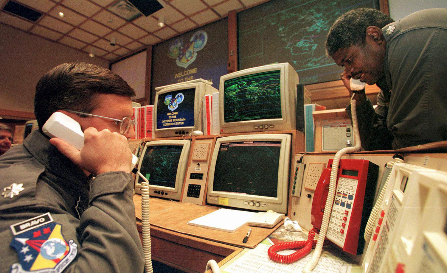 Two men in uniform look worried as they speak on phones at a command center with several phones and computers.