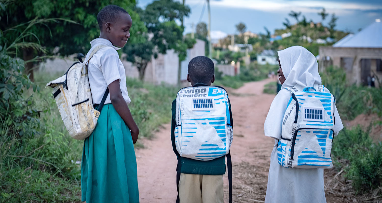 Three children stand on a dirt path wearing Soma backpacks with the solar panels visible.