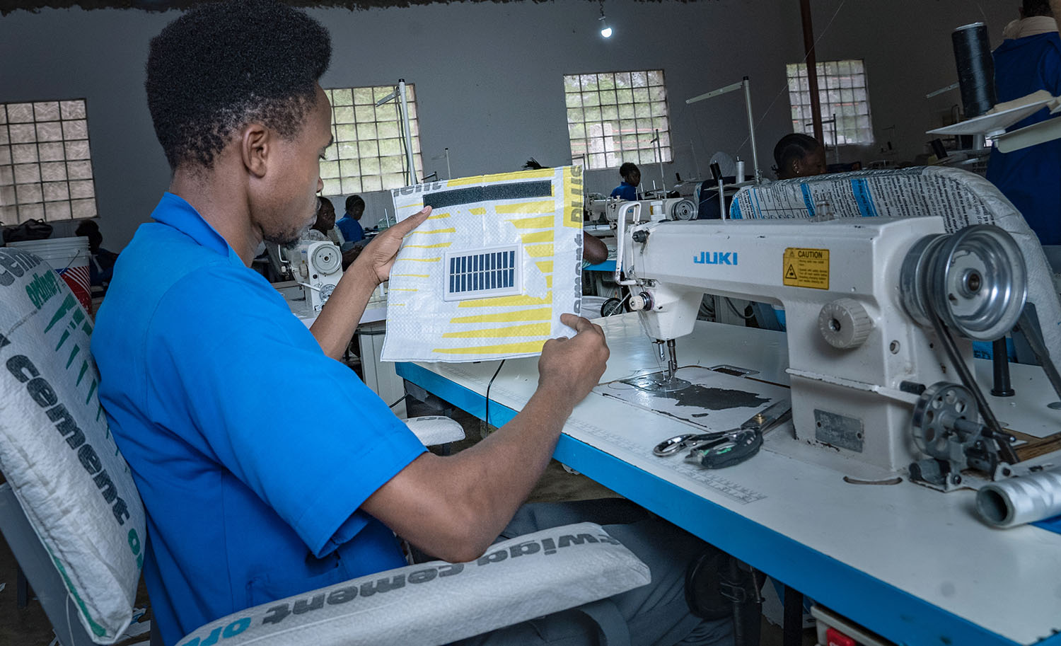 A worker sits at a sewing machine and holds up a small solar panel that has been sewn into material that was once a cement bag.