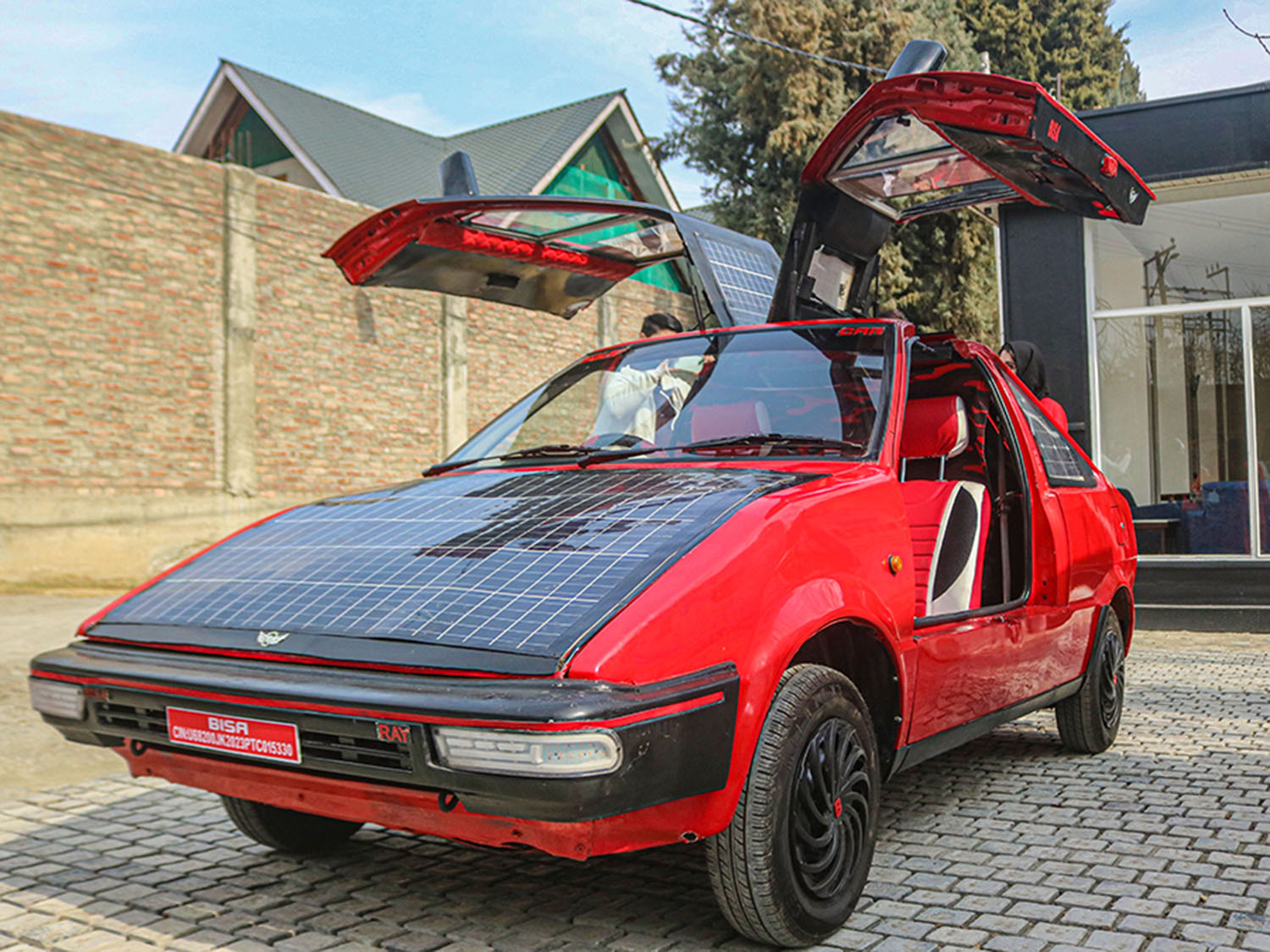 A red car with solar panels on its roof and side rear windows sits in a driveway with its doors open.