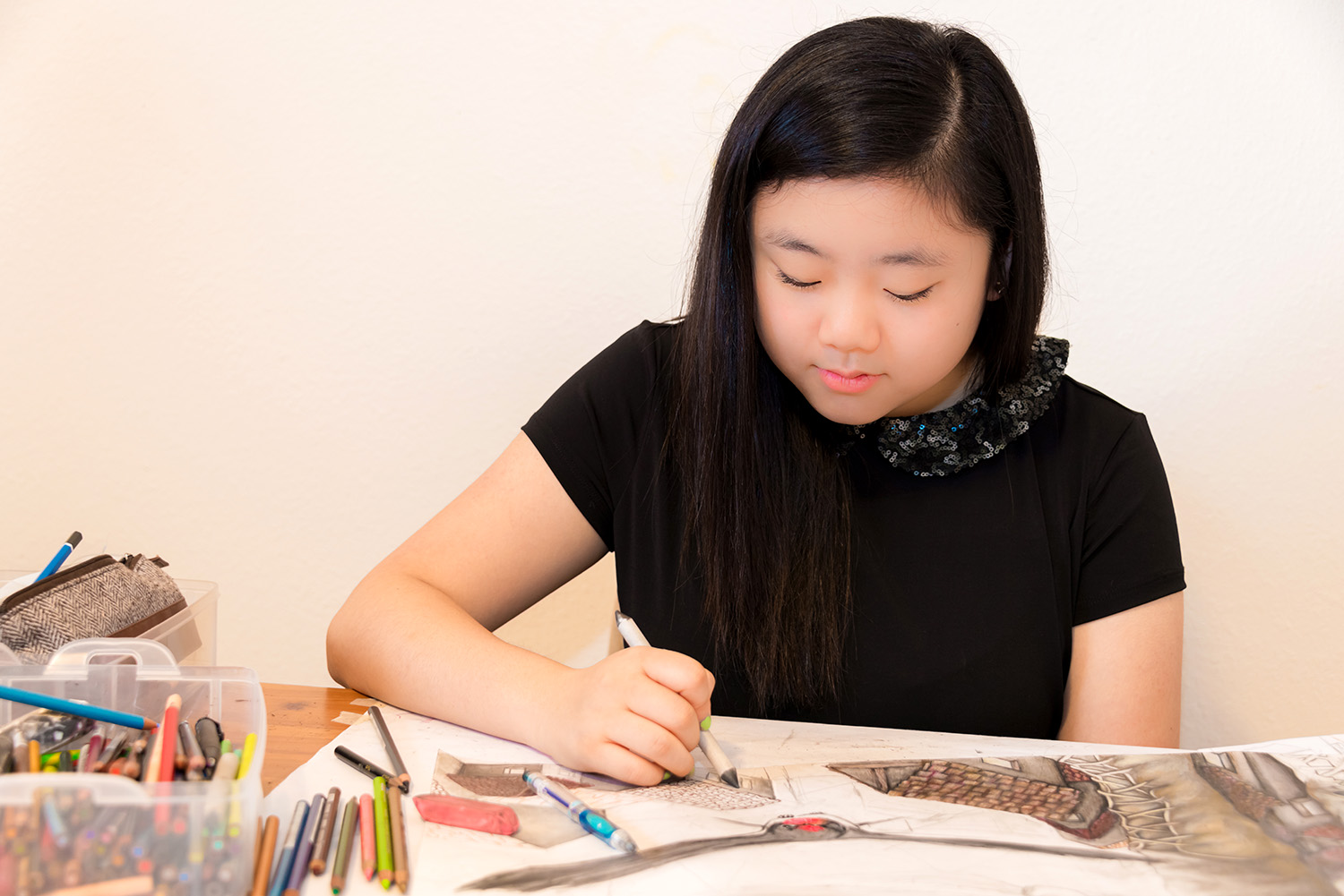 A girl uses a charcoal pencil to draw at a table where there is a container holding many other pencils and other drawing tools.