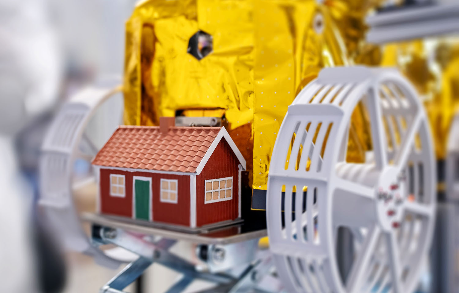 A tiny red house with white trim and a green door sits among some equipment.