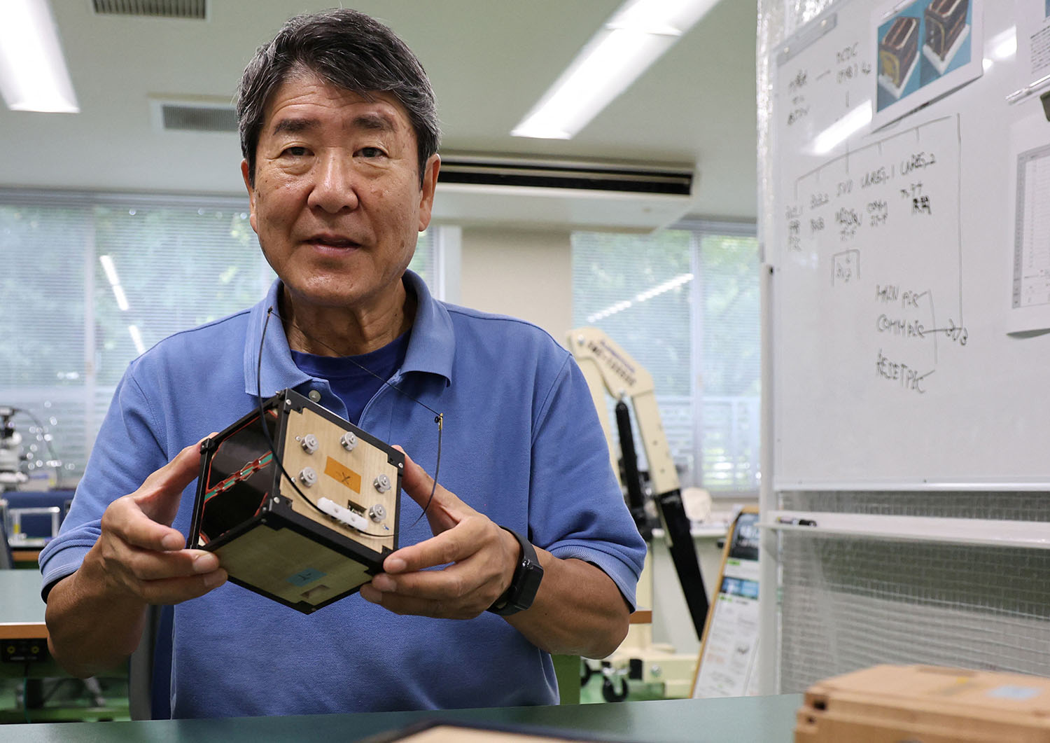 Takao Doi stands in a laboratory holding a small, cube-shaped satellite.