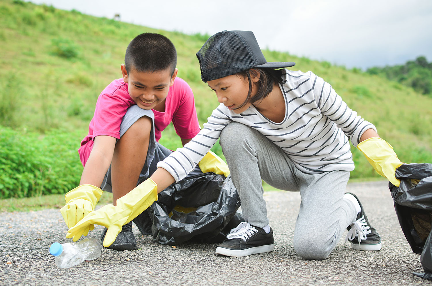 Two children wearing gloves and holding trash bags kneel on pavement and pick up a plastic bottle.
