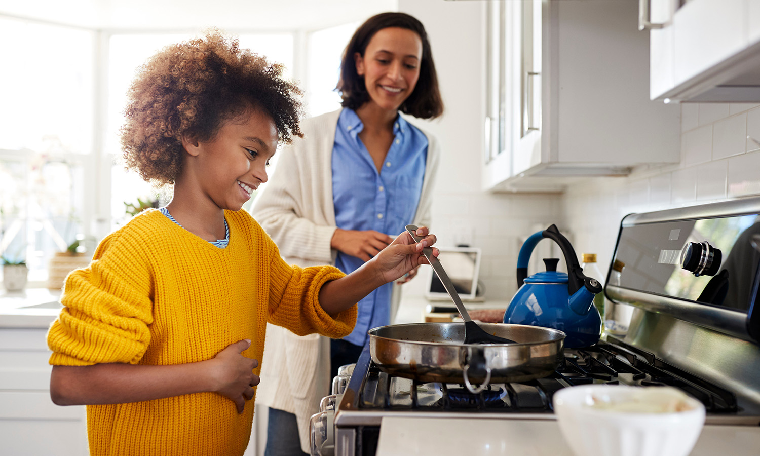 A smiling tween girl uses a spatula in a frying pan while her mother supervises and smiles.