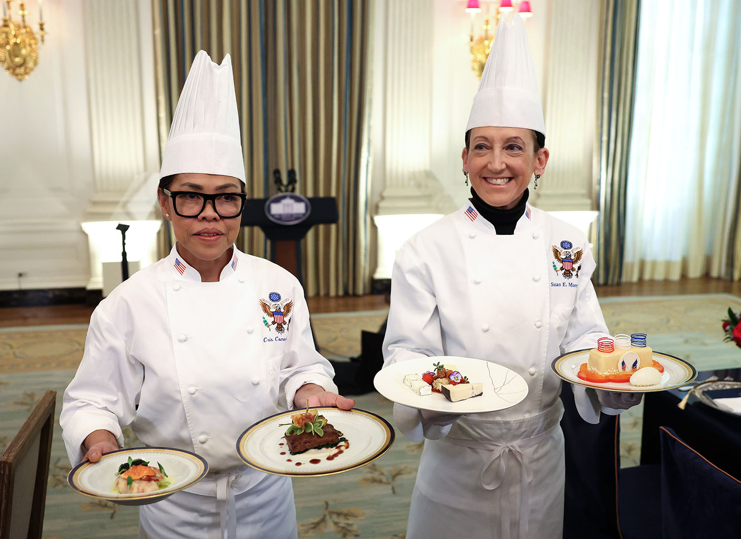 Cristeta Comerford and Susie Morrison stand in a room in the White House wearing chefs’ hats and coats and holding a plate of food in each hand.