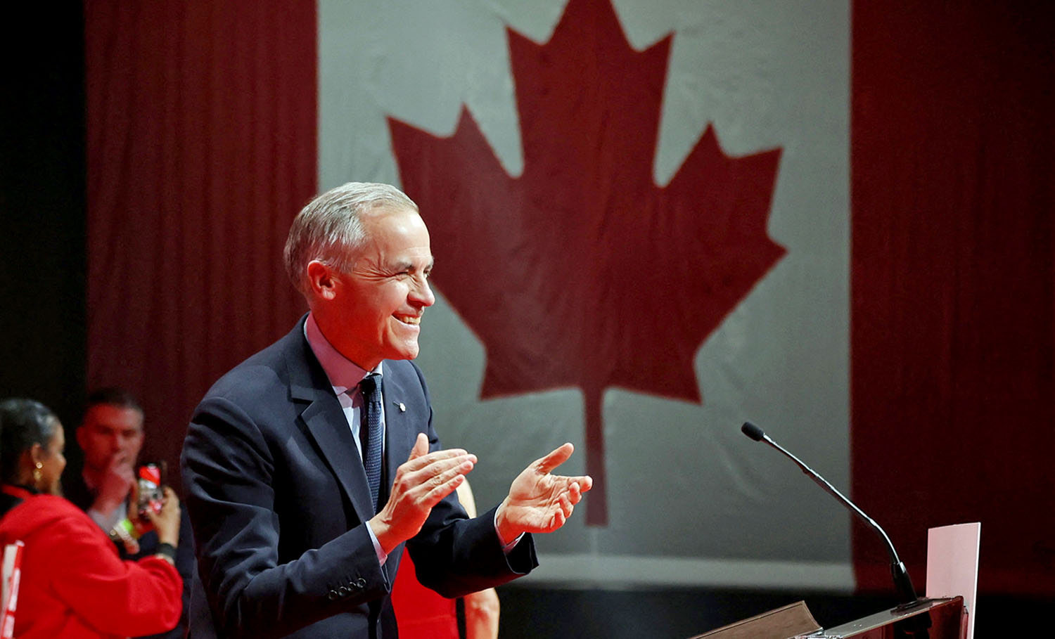 Mark Carney smiles and claps while standing at a podium in front of a Canadian flag.