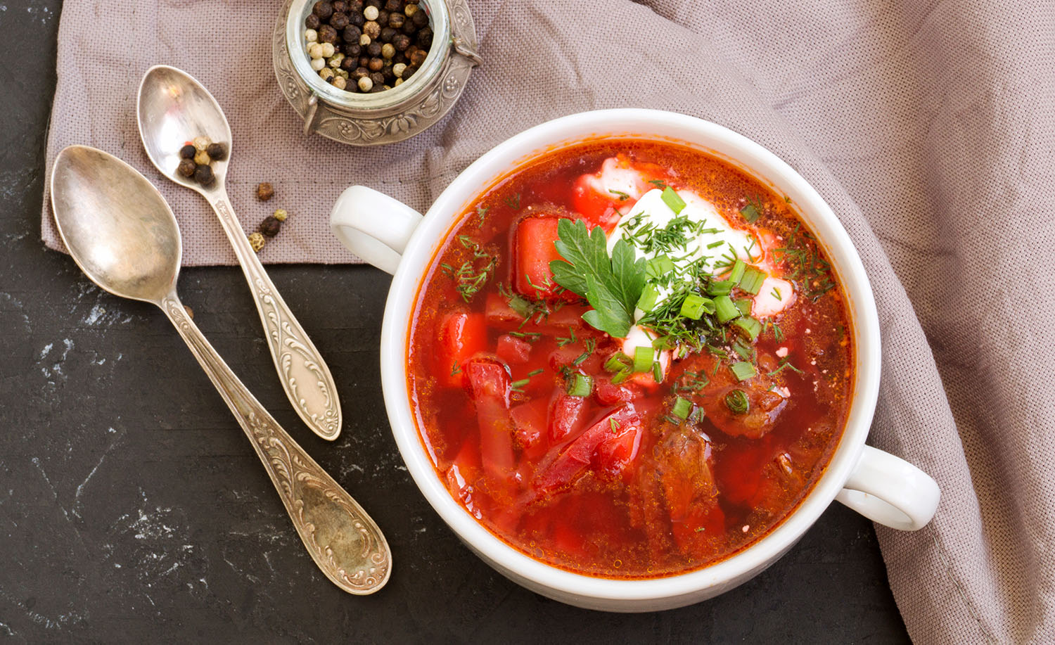 A bowl of red soup containing some large pieces of beet as well as sour cream and chives is next to two spoons.