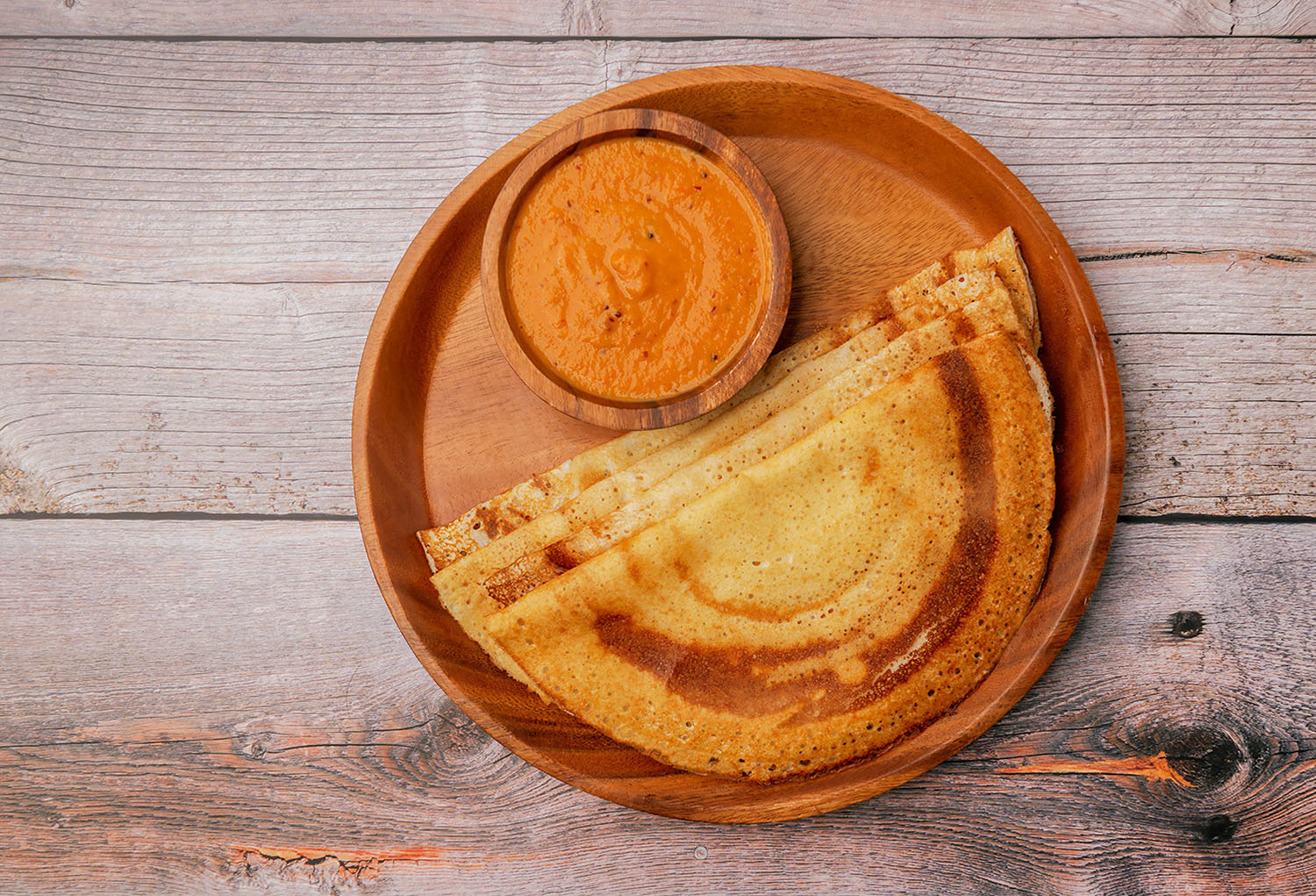 Four thin pancakes on a wooden plate with a small bowl of orange sauce.
