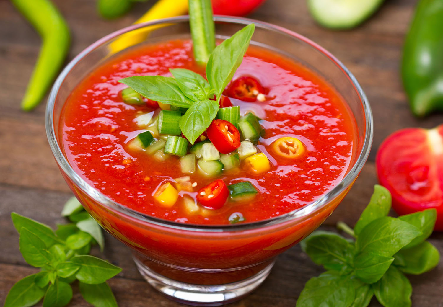 A bowl contains red soup with some chopped green onions, cucumbers, and red peppers.
