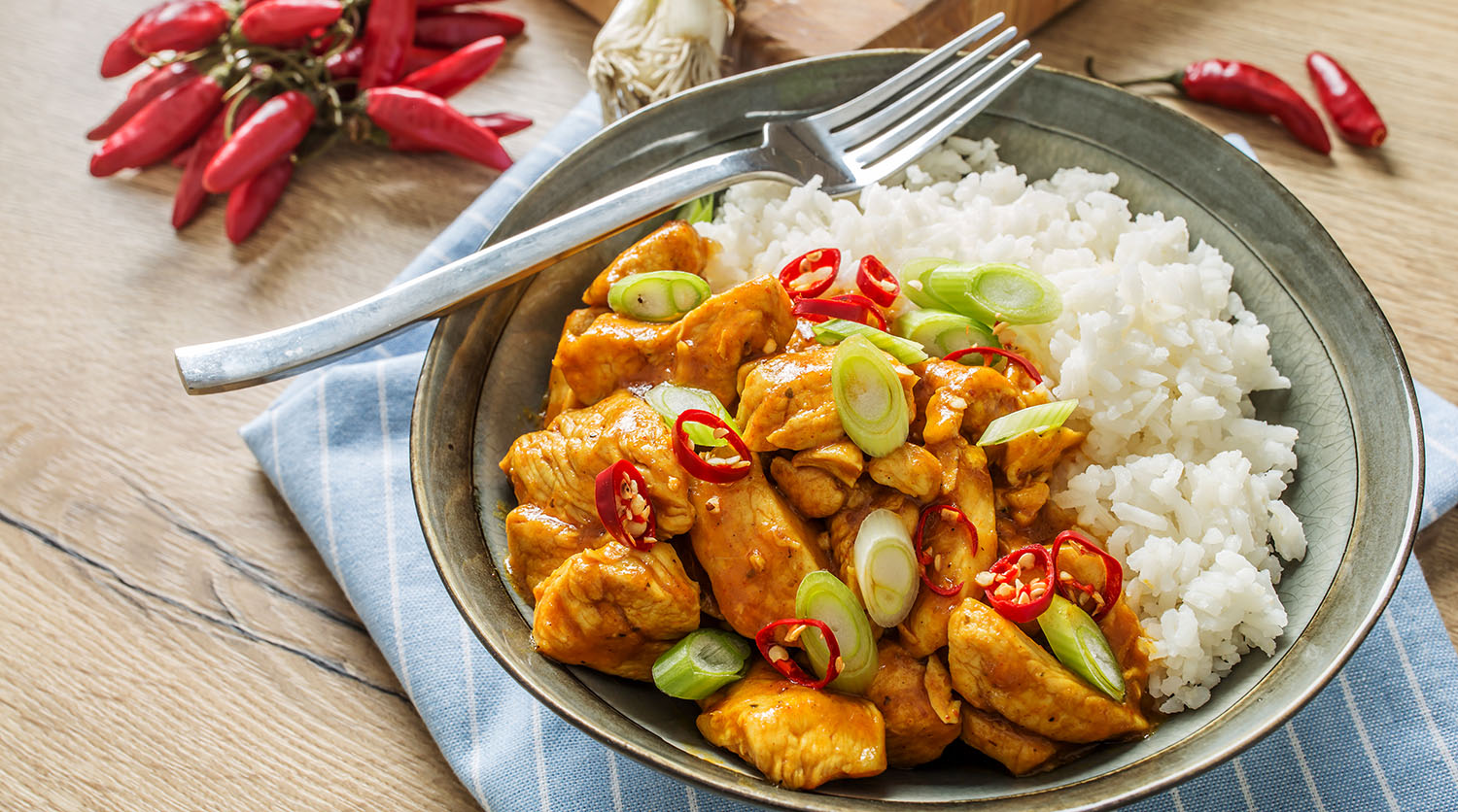 A bowl contains white rice and chicken topped with green onions and red peppers.