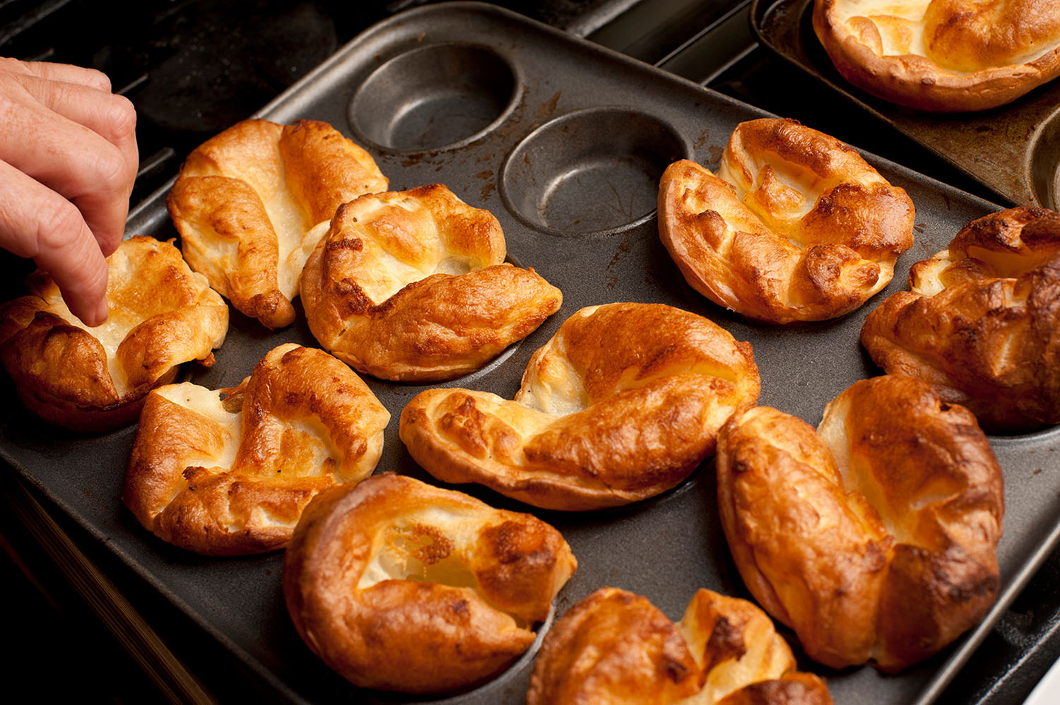 A hand reaches to remove a Yorkshire pudding from a baking tin on which there are 10 Yorkshire puddings.