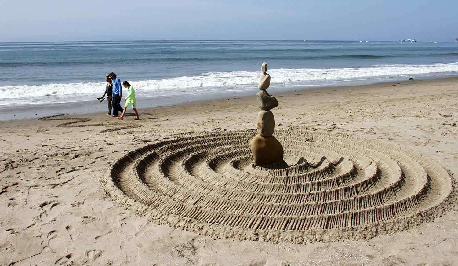 Three people walk on a clean sandy beach on which there is also a stone sculpture.