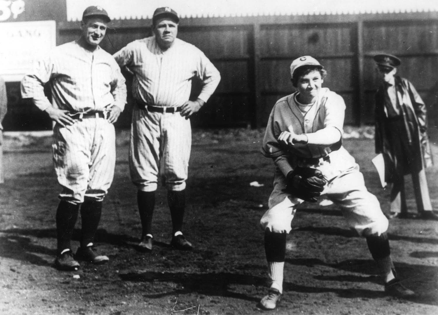 Jackie Mitchell wears a baseball uniform as she pitches while Lou Gehrig and Babe Ruth stand behind and watch.