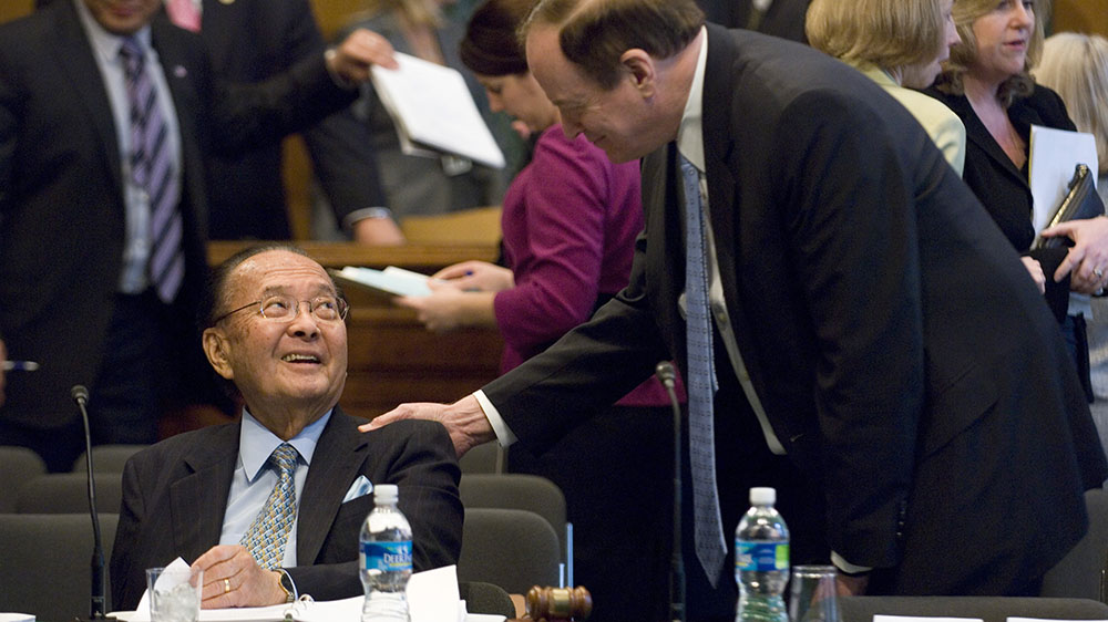 Senator Daniel Inouye is seated and smiles as Senator Richard Shelby greets in with a hand on his shoulder.