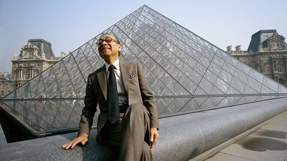 I.M. Pei in a seated pose in front of the glass pyramid addition to the Louvre Museum.