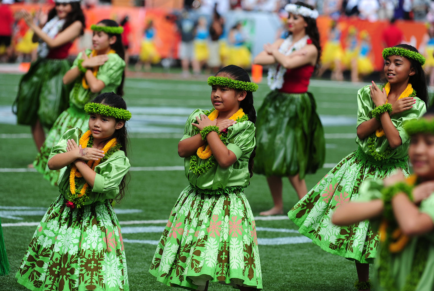 Girls and women perform hula dancing on a sports field while wearing traditional clothing.