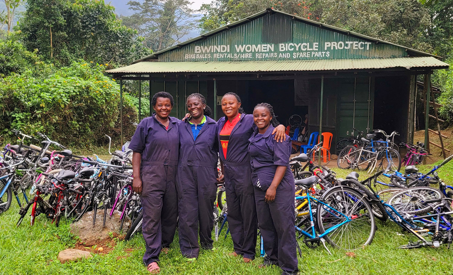 Four women wearing workers’ jumpsuits pose with bicycles outside a shed that reads Bwindi Women Bicycle Project.