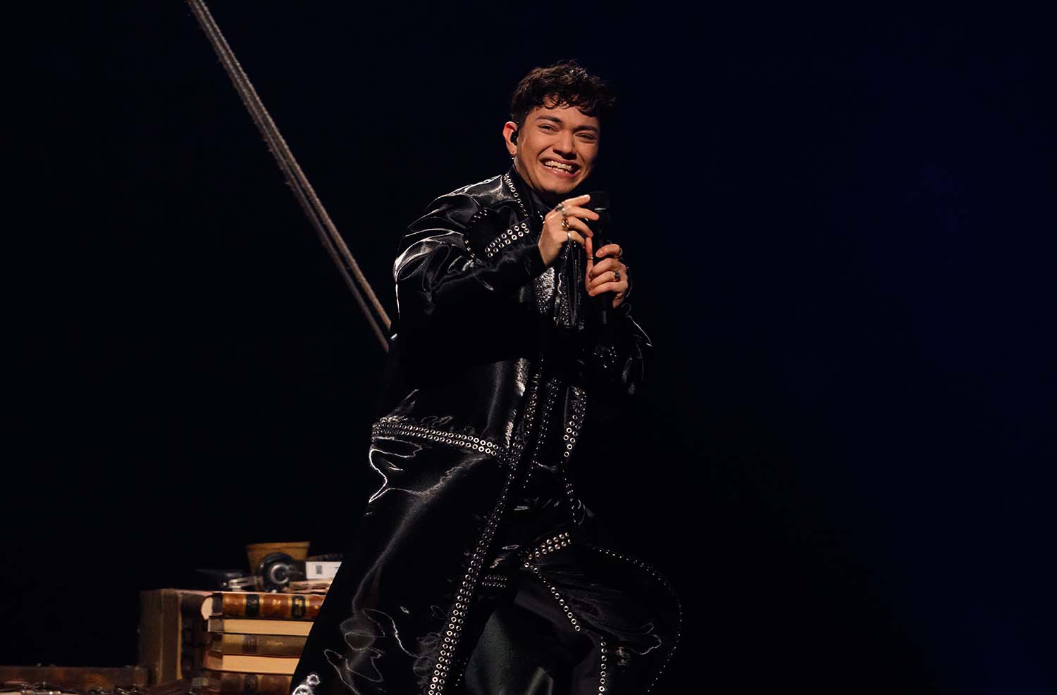 JJ smiles and holds a microphone while wearing a black jumpsuit.