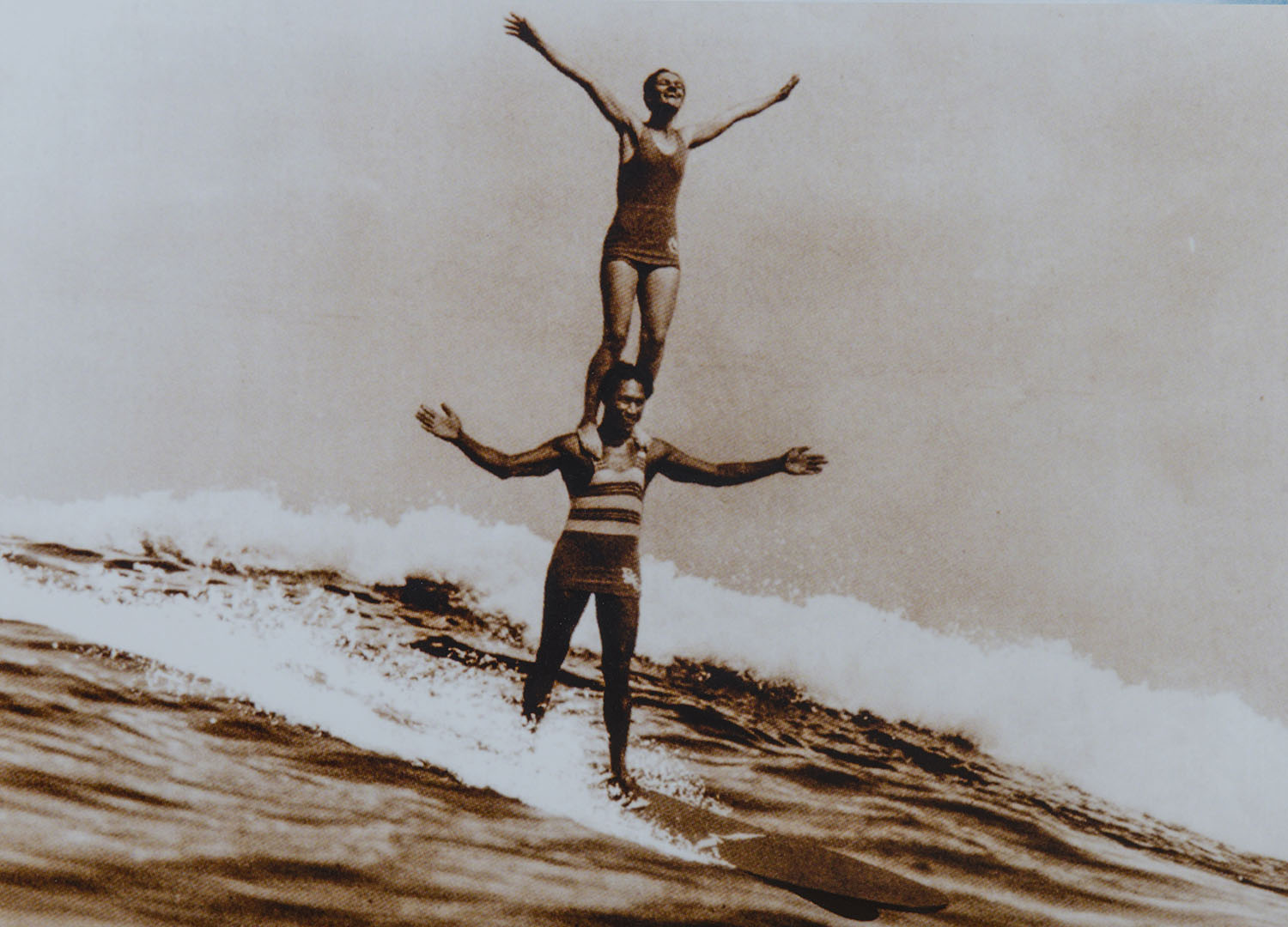 Duke Kahanamoku surfs on a wave with Viola Hartman standing on his shoulders.