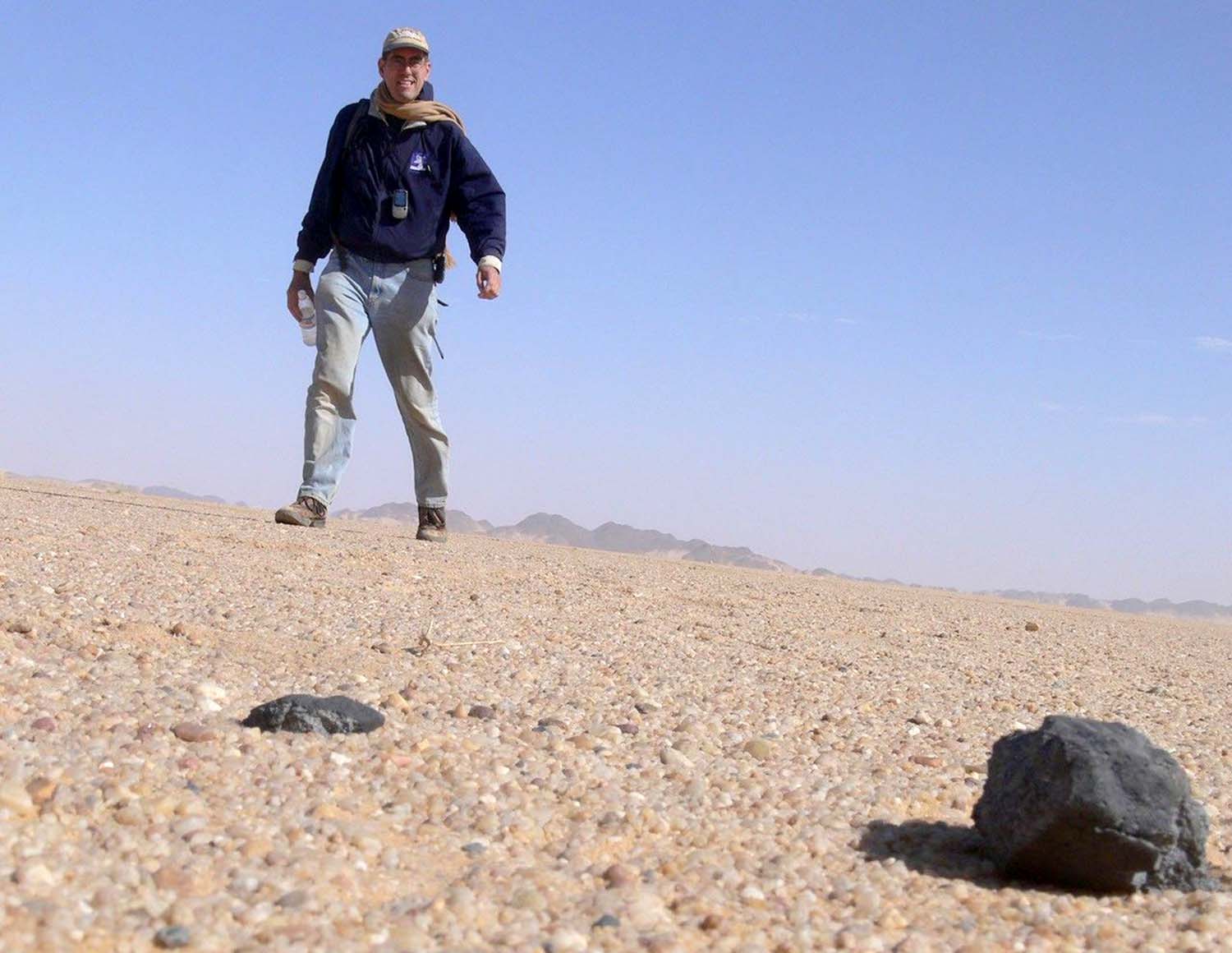 Peter Jenniskens stands in a desert landscape. On the ground in front of him, there are small pieces of meteorites a little larger than his hands.