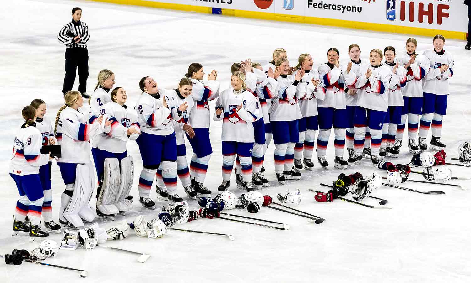 A hockey team made up of young women in their teens is lined up on the ice in uniform with their equipment in front of them