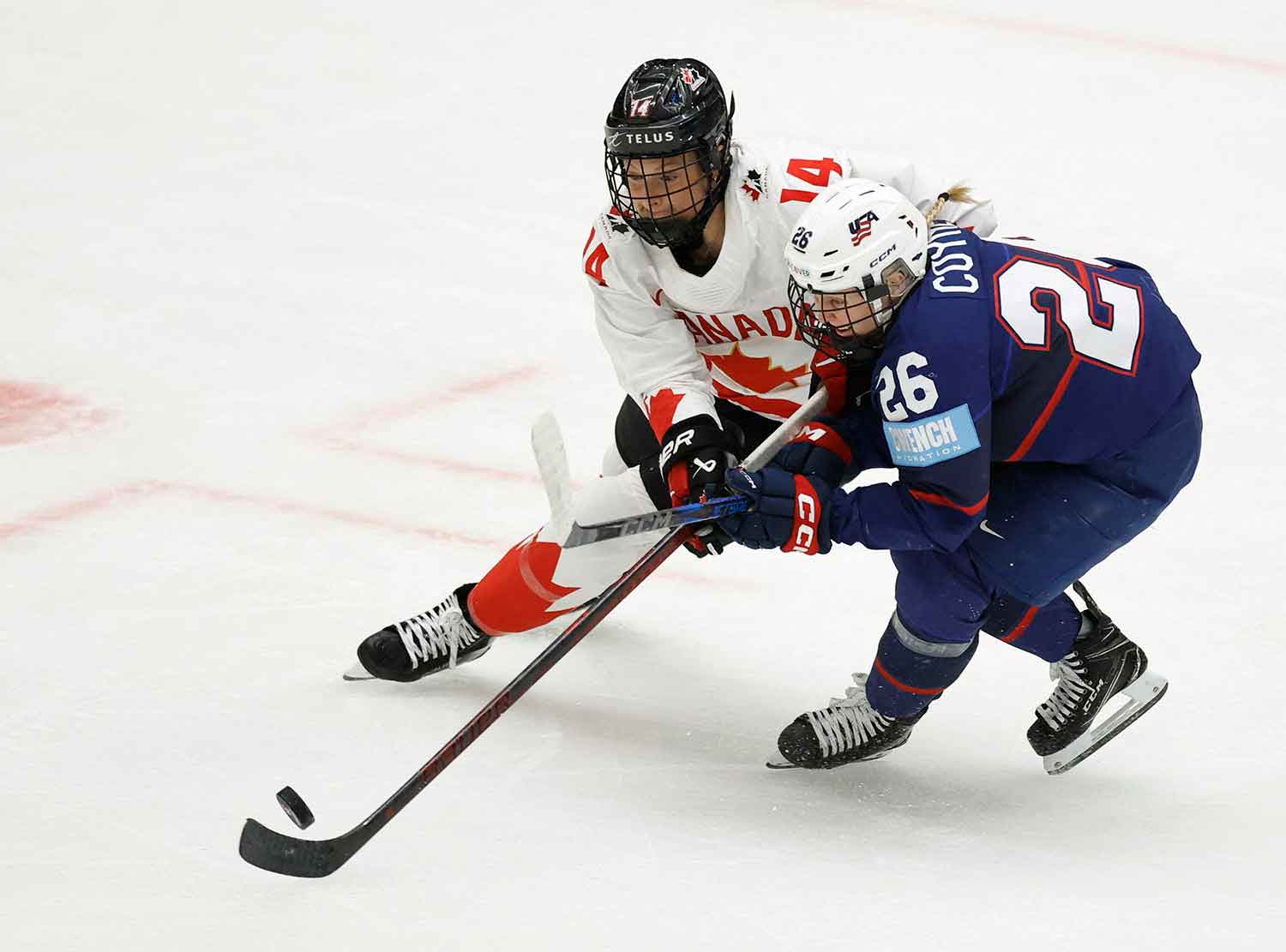 Renata Fast and Kendall Coyne are on the ice and looking at a puck while positioning their hockey sticks to take possession.