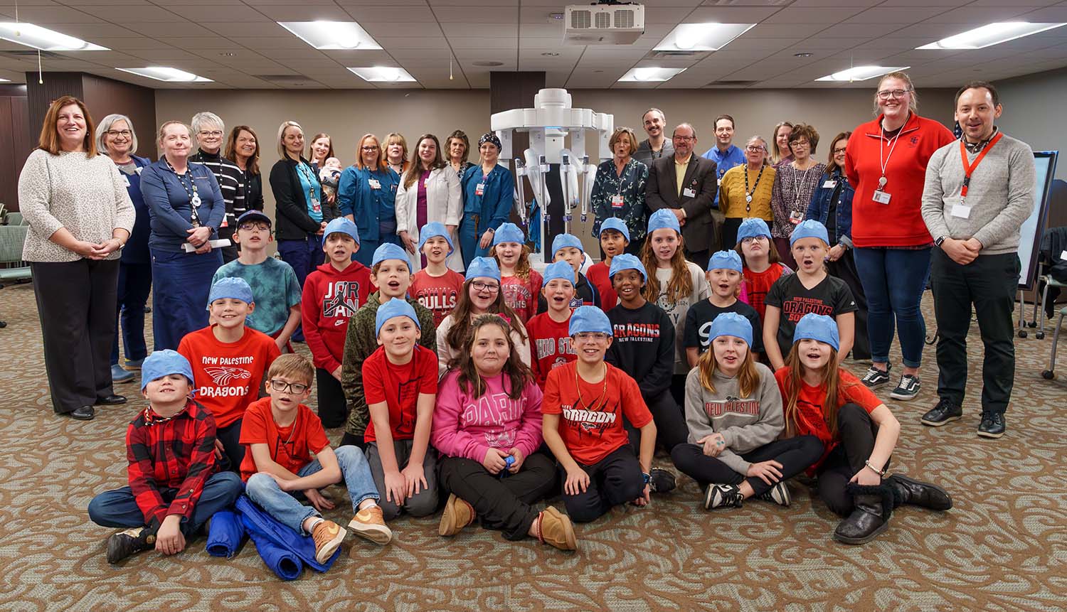 A class of fourth graders sits in front of a large robot that is flanked by men and women wearing hospital ID badges.