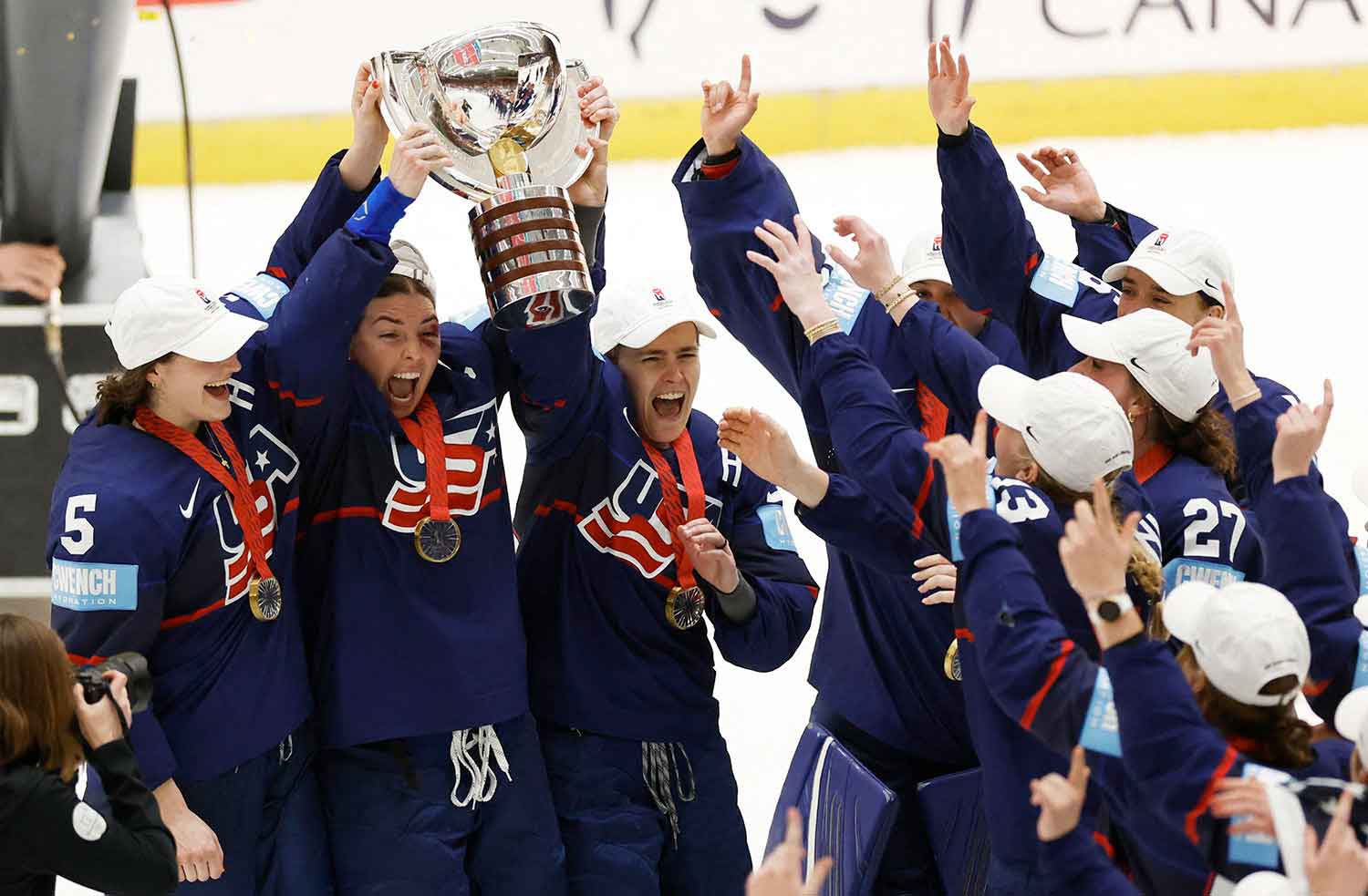 Three hockey players wearing gold medals hold up a trophy as their teammates reach up to touch it.