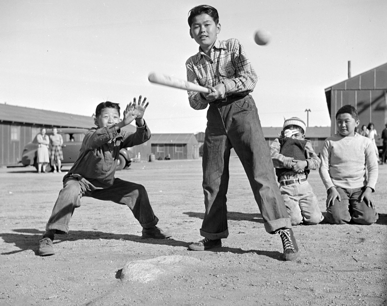 A boy swings a bat at a ball as a catcher is poised behind him, with other children and some adults, as well as barracks, in the background.