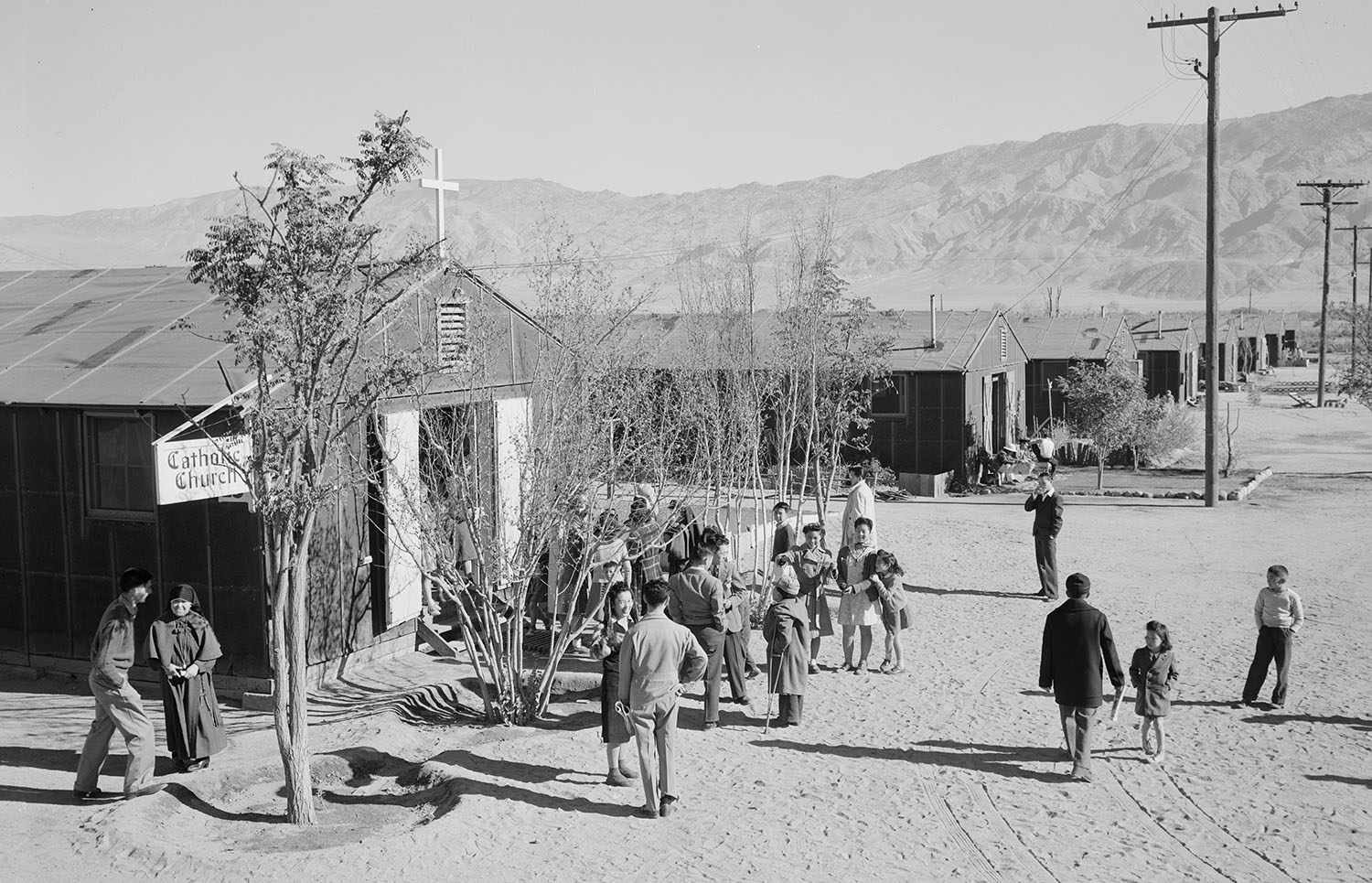 Several adults and some children stand outside a barracks building that has a cross and a sign that says it is a Catholic church.
