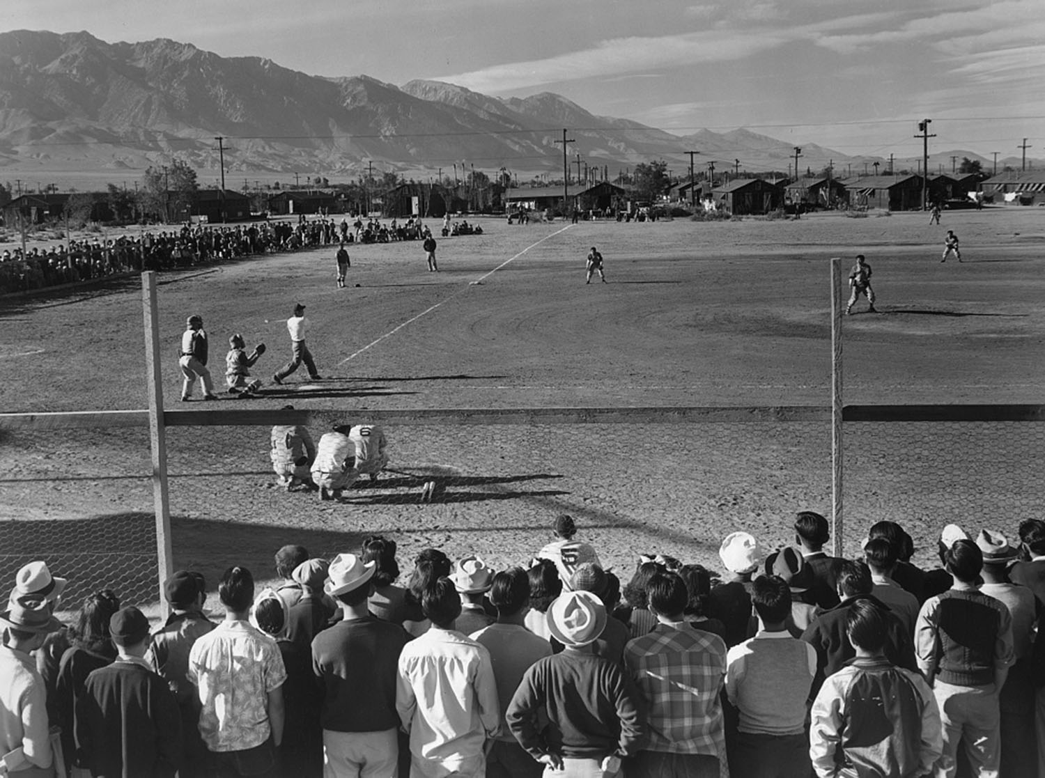 A baseball game is held in front of spectators who are wearing 1940s clothing, with barracks in the background.