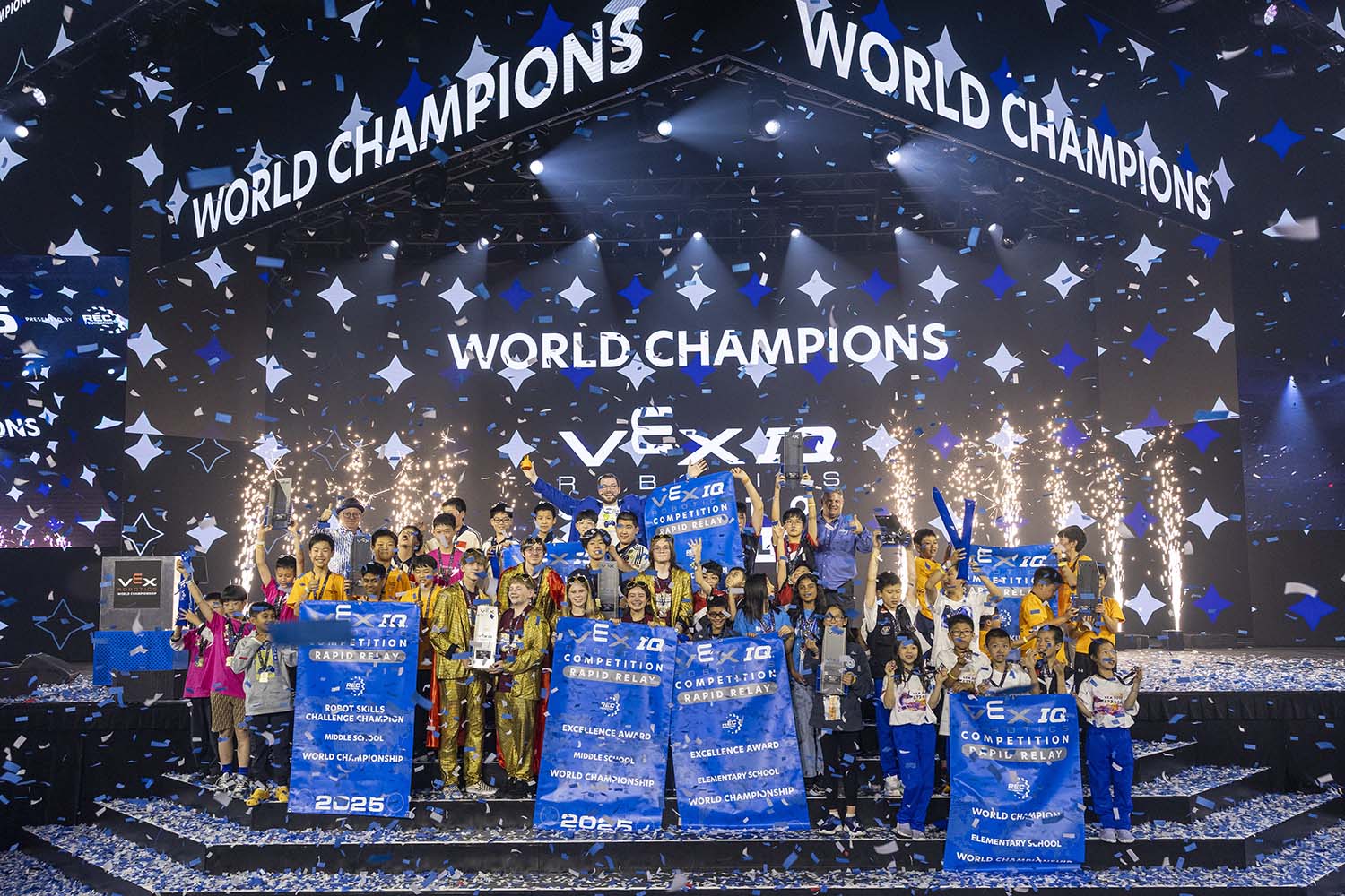 A group of students pose on stage smiling and holding banners that say VEX IQ Competition as confetti falls.