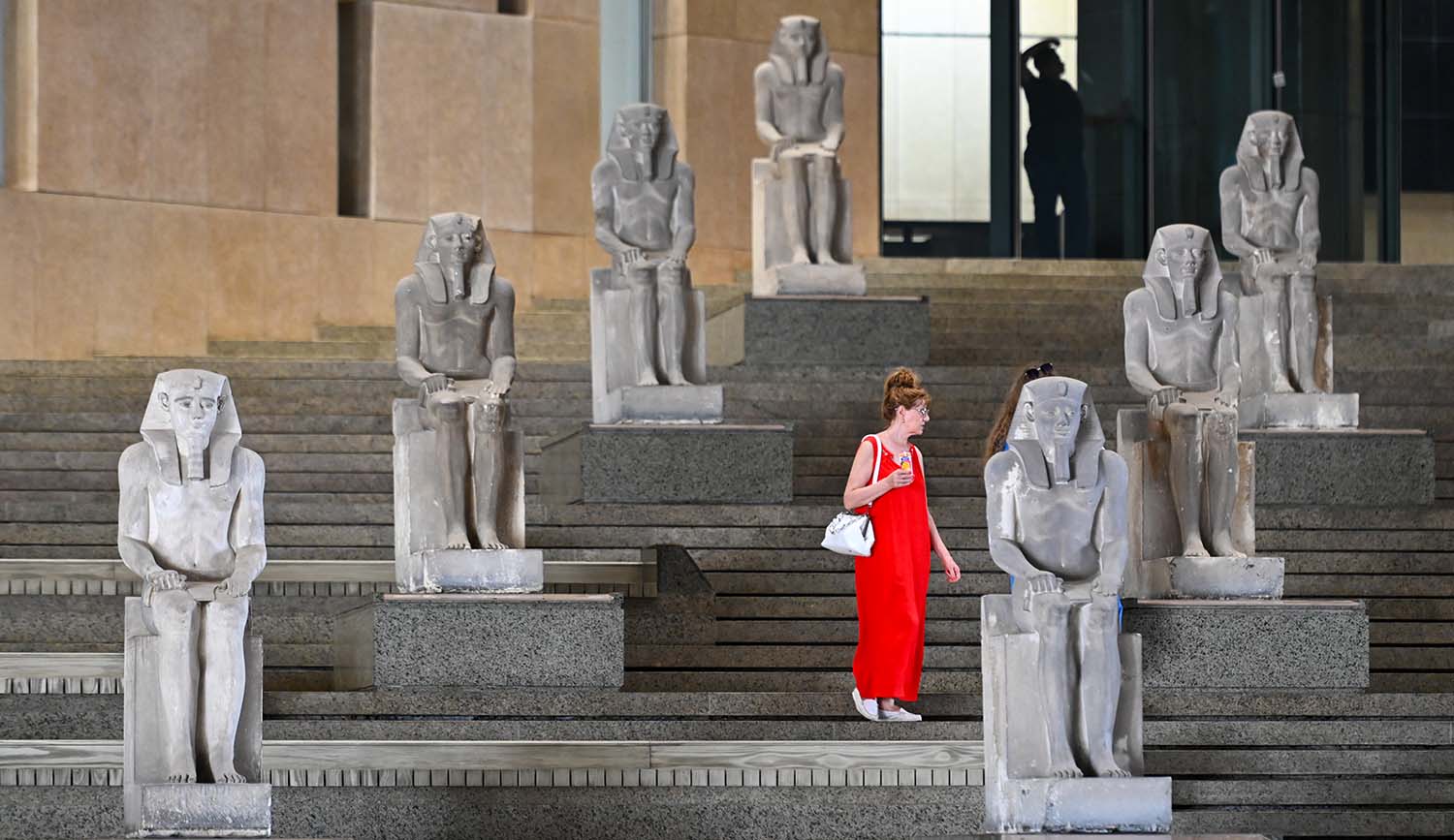 A woman stands on a large staircase on which seven large statues of pharaohs sit.