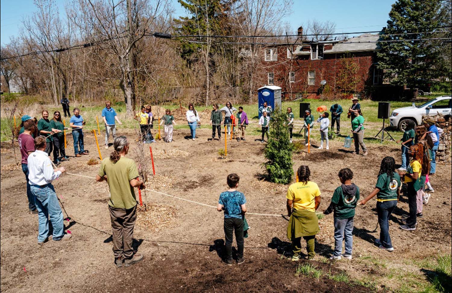 Children and adults stand in a circle around a giant sequoia sapling that has been planted in an empty lot next to a building.